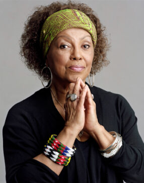 Marta Moreno Vega sitting with her hands in a prayer pose under her chin wearing a green headband with ornate bracelets on her wrist