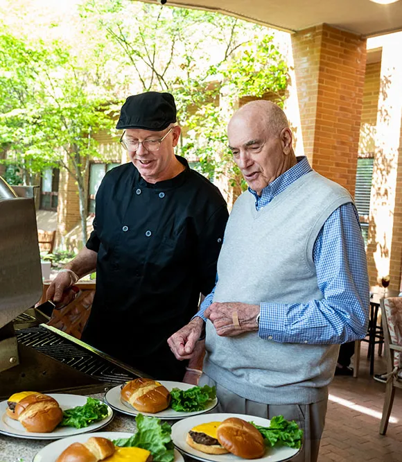 Edenwald_Dining_Catering-Special-Events An older man standing next to a chef grilling hamburgers