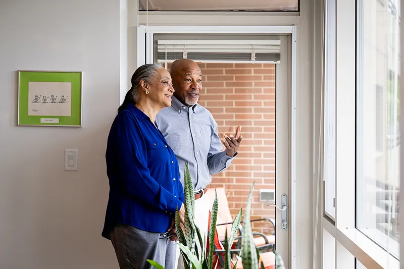 A senior couple standing in their apartment looking out the window